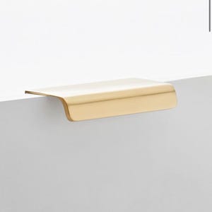 Puede incluir: Un tirador de armario de color dorado con un borde curvo. El tirador está hecho de metal y tiene un acabado liso y pulido.