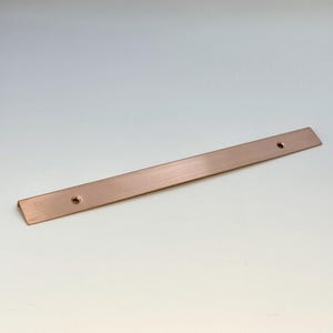 May include: A copper-coloured metal bar with two pre-drilled holes. The bar is rectangular with a slightly bent end. The surface has a brushed texture. It is likely a furniture or cabinet hardware component.