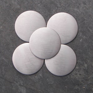 May include: Five silver-coloured, brushed metal circles arranged in a flower-like pattern. Each circle has a textured, linear grain. The arrangement is centered on a dark grey, textured surface.