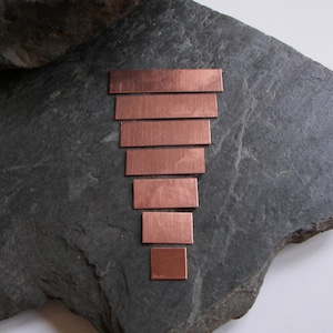 May include: A set of seven copper metal strips arranged in a graduated size, forming a pyramid shape. The strips are on a dark gray stone surface.