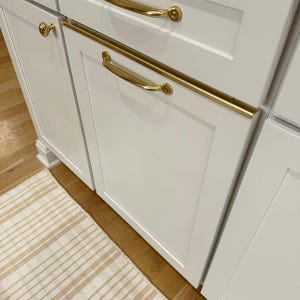 May include: Close-up of white kitchen cabinets with gold-colored hardware. The cabinets have a recessed panel design and are accented with long, curved handles and round knobs. A striped rug in neutral tones is visible in the foreground.