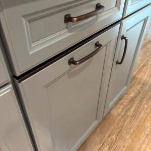 May include: Close-up of a section of light gray kitchen cabinets with bronze-colored hardware. The cabinets have a recessed panel design and are fitted with curved bar pulls and a single handle. The hardware provides a contrast to the light gray color of the cabinets.