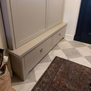 May include: A light beige cabinet with two drawers and gold-colored handles. The cabinet is part of a larger unit and sits on a tiled floor. A patterned rug is in front of the cabinet.