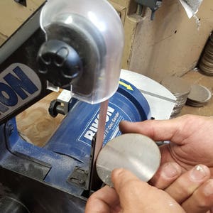 May include: A blue and black disc sander with a clear plastic guard. A person is holding a silver metal disc against the sander.