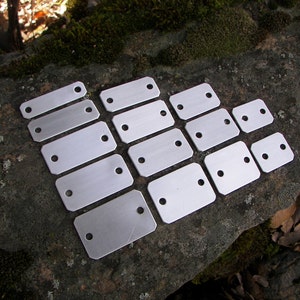 May include: A set of 14 silver metal tags with two holes each. The tags are arranged in a staggered pattern on a gray, textured surface.