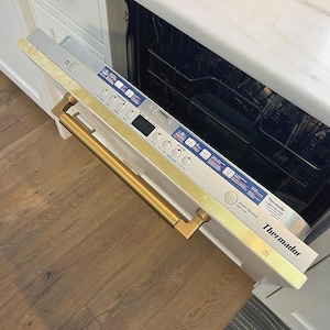 Puede incluir: Un lavavajillas de acero inoxidable con un tirador y molduras doradas. El panel de control tiene varios botones y una pantalla digital. La marca "Thermador" es visible.