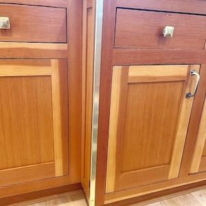 May include: Wooden kitchen cabinets with a natural finish. The cabinets have drawers and doors with square knobs and handles. A vertical metal strip with a gold finish runs along the edge of the cabinets.