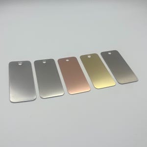 May include: Five rectangular metal tags in varying colors: silver, gray, rose gold, gold, and brushed silver. Each tag has a rounded top with a small hole for hanging. The tags are arranged in a row.