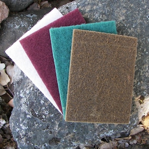 May include: Four different colored scouring pads, one white, one maroon, one teal, and one brown, are arranged on a gray rock surface.