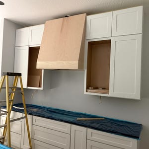 Könnte beinhalten: Eine Kücheninstallation mit weißen Schränken im Shaker-Stil mit flachen Türen und Schubladen. Eine große, unbehandelte Dunstabzugshaube aus hellem Holz ist mittig zwischen den Oberschränken platziert. Die Unterschränke sind mit blauer Plastikfolie abgedeckt.
