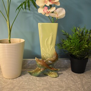 Puede incluir: Un jarrón de cerámica verde claro con un diseño floral estilizado y una base de pájaro marrón y verde. El jarrón está lleno de orquídeas blancas con acentos rosados.