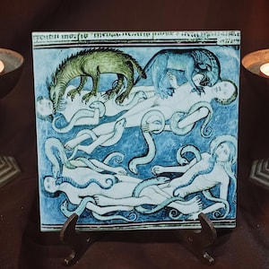 Könnte beinhalten: Eine quadratische, blau-weiße Fliese mit einer Illustration im mittelalterlichen Stil von Figuren und Kreaturen. Ein grüner Drache, ein blauer Löwe und Figuren mit schlangenartigen Anhängseln sind dargestellt. Zwei brennende Kerzen in Bronzehaltern flankieren die Fliese.