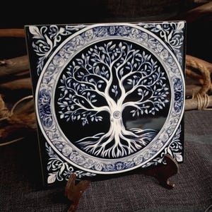 Puede incluir: Azulejo cerámico cuadrado con un diseño de árbol de la vida blanco sobre fondo negro, enmarcado por un borde decorativo con detalles azules. El azulejo se exhibe sobre un pequeño soporte marrón, mostrando sus intrincados detalles.