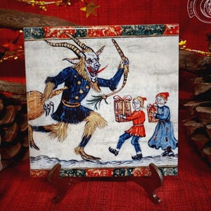 Puede incluir: Azulejo de cerámica cuadrado con una ilustración de Krampus. La imagen representa una figura con cuernos con un látigo y dos niños que llevan regalos. El azulejo tiene un borde decorativo y se exhibe en un soporte.