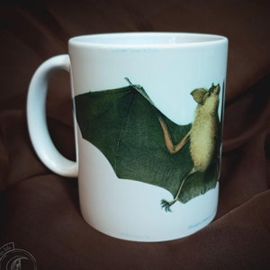 Może przedstawiać: Biały kubek ceramiczny z ilustracją brązowego nietoperza. Nietoperz ma zielone skrzydła i jest przedstawiony w locie.
