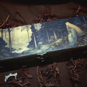 Puede incluir: Una caja de madera con tapa de bisagras, que presenta una escena pintada de una mujer de pie en un bosque. La mujer tiene el pelo largo y suelto y lleva un vestido blanco. El bosque está representado en tonos verdes y marrones, con un camino que se adentra en la distancia.