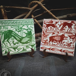 May include: Two ceramic tiles with hand-painted designs. The tile on the left has a green and white design of a mythical creature with wings. The tile on the right has a red and white design of a mythical creature with wings.