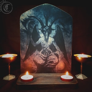 Puede incluir: Un altar de madera oscura con cuatro velas, dos a cada lado de una imagen enmarcada de una figura con cuernos y alas. La imagen está sobre un fondo azul oscuro y rojo.