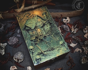 May include: A wooden box featuring a green and gold illustration of a house on stilts within a forest setting. The house has a pointed roof and windows. The forest is filled with trees and vines. Skulls are scattered throughout the forest. The box is closed.