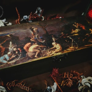 Op de afbeelding: Een houten doos met een scharnierend deksel, met een geschilderd tafereel van mensen die rond een vuur zijn verzameld. De doos heeft een donkerbruine afwerking en een metalen sluiting.