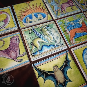 May include: A set of square ceramic tiles, each adorned with medieval-style illustrations. The tiles showcase various designs, such as a griffin, lion, and bat, painted in vivid colours on a yellow-green backdrop. The artwork is framed with decorative borders, reminiscent of illuminated manuscripts.