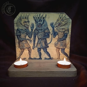 May include: Wooden candle holder featuring a painted panel with three demonic figures in blue, tan, and gold. The figures have horns and tails. Two lit tea light candles are placed in front of the panel.