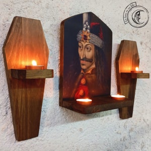 May include: Three wooden coffin-shaped candle holders with shelves, each with a lit candle. The centre holder has a framed portrait of Vlad the Impaler, a historical figure associated with vampires, with the text "La luna en un hilo" above the image.