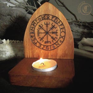 Könnte beinhalten: Kerzenhalter aus Holz mit einem geschnitzten Vegvisir-Symbol. Der Kerzenhalter ist aus dunklem Holz gefertigt und hat eine brennende Teelichtkerze. Das Vegvisir-Symbol ist ein Wikingerkompass, ein Symbol für Schutz und Führung.