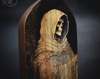 May include: A wooden plaque featuring a painted image of a skeletal figure wearing a white hooded robe. The figure is depicted with a skull face and is set against a dark background.