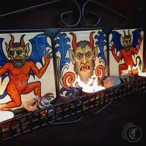 May include: A decorative candle holder featuring three panels with devilish figures in red, orange, and blue. The metal frame holds candles and decorative elements. The artwork has a vintage, gothic style.