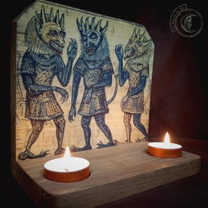 K&ouml;nnte beinhalten: Holzkerzenhalter mit einer geschnitzten Illustration von drei d&auml;monischen Figuren in Blau und Beige. Zwei brennende Teelichter befinden sich vorne. Das Kunstwerk befindet sich auf einer hellen Holztafel.