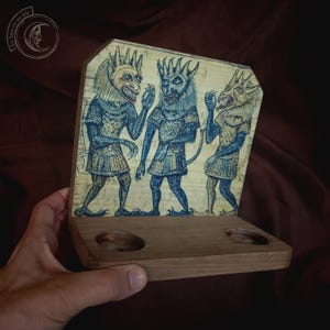 K&ouml;nnte beinhalten: Dekoratives Holzst&uuml;ck mit drei blau get&ouml;nten Figuren mit geh&ouml;rnten Masken und mittelalterlicher Kleidung. Das Kunstwerk ist auf einem Holzsockel mit zwei kreisf&ouml;rmigen Aussparungen montiert. Das St&uuml;ck hat eine Vintage-&Auml;sthetik.