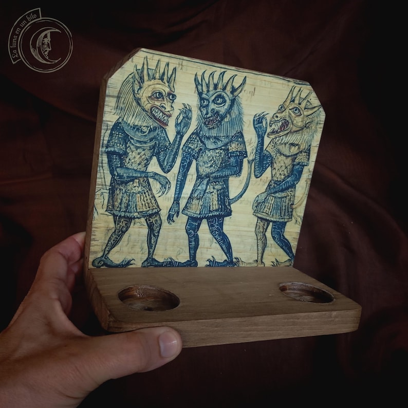 K&ouml;nnte beinhalten: Dekoratives Holzst&uuml;ck mit drei blau get&ouml;nten Figuren mit geh&ouml;rnten Masken und mittelalterlicher Kleidung. Das Kunstwerk ist auf einem Holzsockel mit zwei kreisf&ouml;rmigen Aussparungen montiert. Das St&uuml;ck hat eine Vintage-&Auml;sthetik.