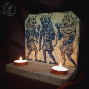 K&ouml;nnte beinhalten: Holzkerzenhalter mit einer mittelalterlichen Illustration von drei d&auml;monischen Figuren in blauer und beiger Kleidung mit geh&ouml;rnten K&ouml;pfen. Zwei brennende Teelichter befinden sich vor der Illustration. Der Text "La luna en un hilo" befindet sich in der oberen linken Ecke.