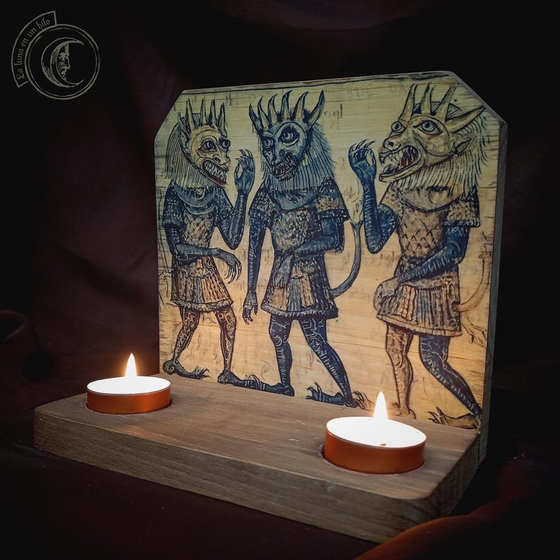 K&ouml;nnte beinhalten: Holzkerzenhalter mit einer mittelalterlichen Illustration von drei d&auml;monischen Figuren in blauer und beiger Kleidung mit geh&ouml;rnten K&ouml;pfen. Zwei brennende Teelichter befinden sich vor der Illustration. Der Text "La luna en un hilo" befindet sich in der oberen linken Ecke.