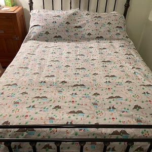 May include: A bed with a patterned quilt and pillowcase set. The bedding features a repeating design of mountains, trees, and small houses in shades of brown, green, red, and blue on a light beige background. The bed frame is black.
