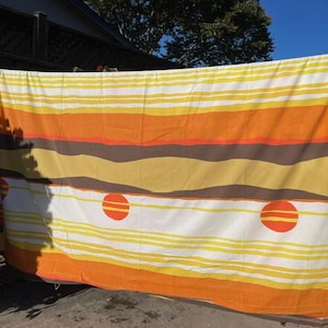 May include: A rectangular fabric panel featuring horizontal stripes in shades of yellow, orange, and brown. The design includes a white section with thin yellow lines and two prominent orange circles, each containing two horizontal yellow stripes.