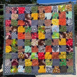 May include: A square patchwork quilt featuring a grid of multi-colored fabric squares with various floral, polka dot, and geometric patterns. The quilt is tied with dark navy blue yarn knots at the intersections and finished with a dark blue border.