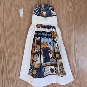 May include: A hanging kitchen towel with a cat-themed patchwork design. The towel features a cream-colored base with a patterned top and a central panel with cat illustrations, hearts, and birdhouses. The text "a cat's a cat that's that" is visible.