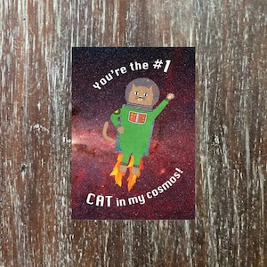 Könnte beinhalten: Eine Grußkarte mit einer Comic-Katze im Astronautenkostüm in einem grünen Raumanzug, die durch den Weltraum fliegt. Die Karte lautet "You're the #1 CAT in my cosmos!"