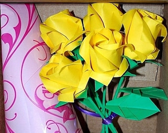 May include: A bouquet of five yellow origami roses with green stems and leaves, tied with a purple ribbon, in a pink and white patterned paper wrapper.