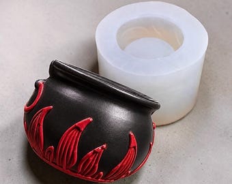 May include: A black cauldron with raised red flame-like details along the base and a translucent white silicone mold positioned behind it. The cauldron features a wide rim and a rounded body, while the mold is cylindrical with a hollow center.