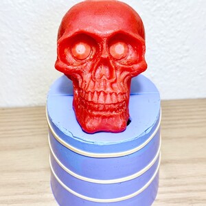 May include: A red skull-shaped candle with white eyes sits on top of a purple silicone mold. The mold has three white bands around it.