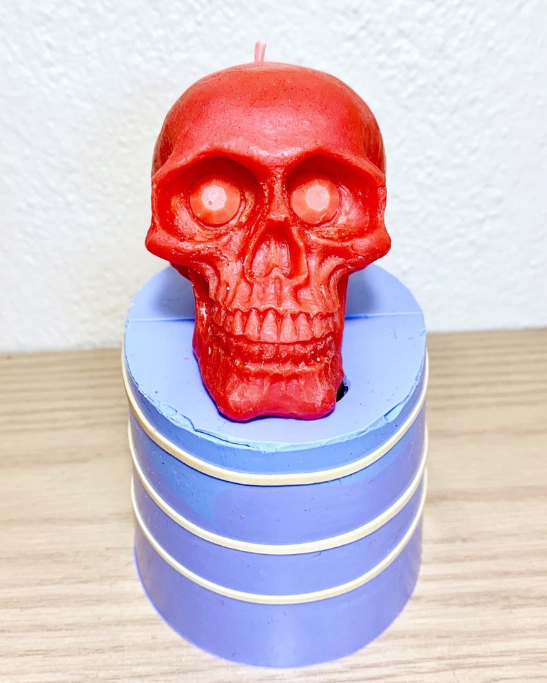 May include: A red skull-shaped candle with white eyes sits on top of a purple silicone mold. The mold has three white bands around it.