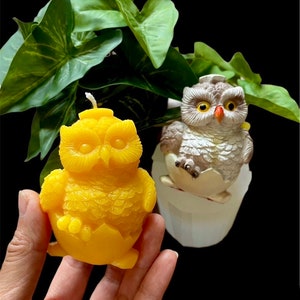 May include: Two owl candles, one yellow and one gray, are shown in the image. The yellow owl candle is in a hand, and the gray owl candle is in a white mold. Both owls are in a hatching position, with their wings spread out.