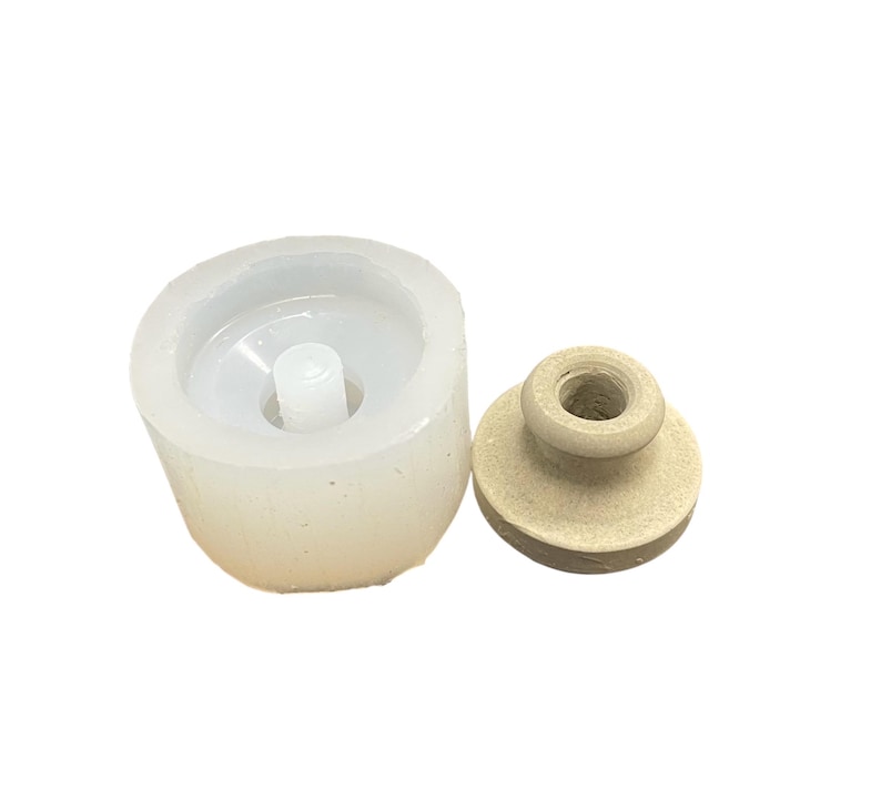May include: A white silicone mold for making a small bottle with a wooden stopper. The mold is round and has a small hole in the center for the stopper.