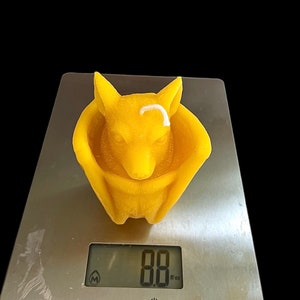May include: A yellow beeswax candle shaped like a fox curled up in a nest. The candle is on a digital scale that reads 8.8 oz.