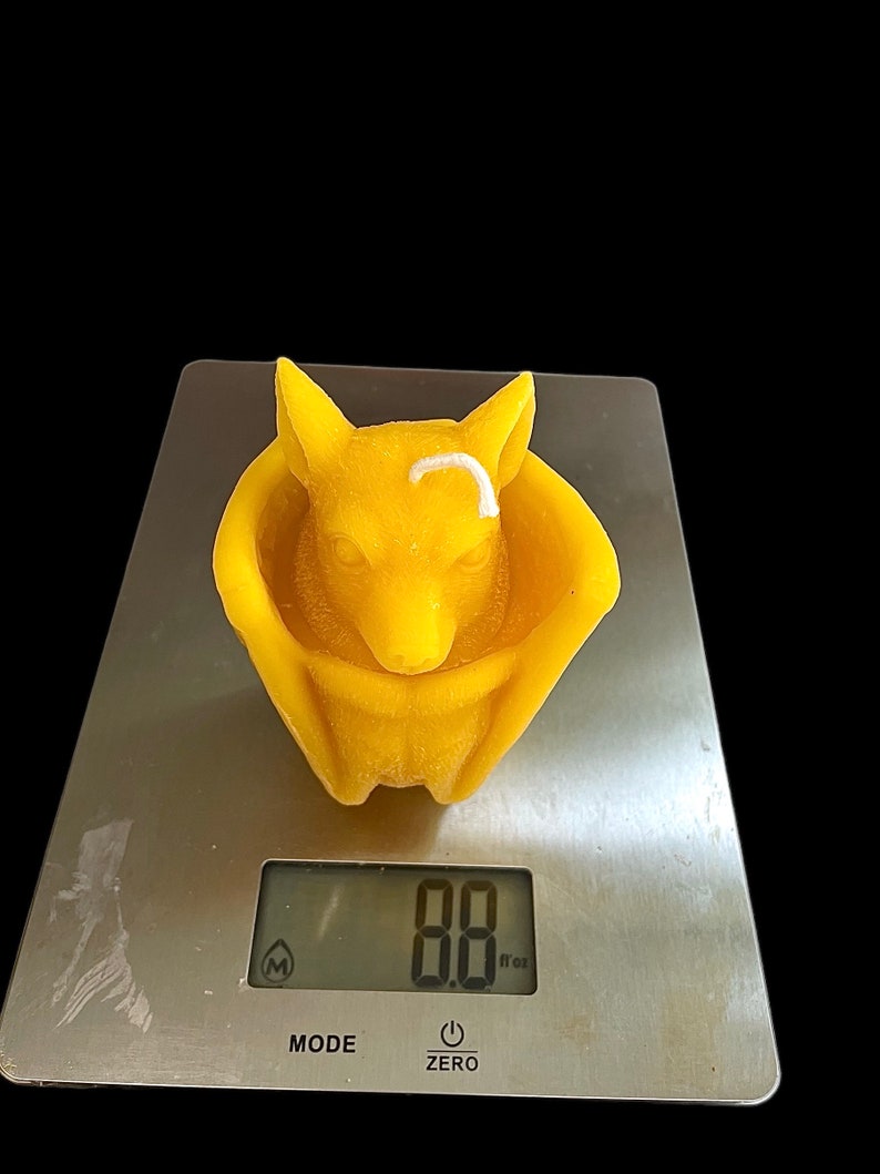 May include: A yellow beeswax candle shaped like a fox curled up in a nest. The candle is on a digital scale that reads 8.8 oz.