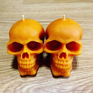 May include: Two orange skull-shaped candles with a white wick on top. The candles are sitting on a wooden surface.