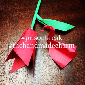 May include: Two origami figures, a red rose and a red bird, are folded from paper. The rose is partially folded and the bird is fully folded. The figures are on a wooden surface. The text #prisonbreak #thehandmadecharm is written on the surface.